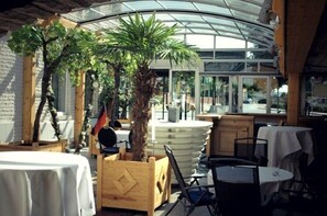 An indoor dining area with tables and chairs, a large central plant, and a glass ceiling. - Hotel & Restaurant Bei Baki (Sehnde)