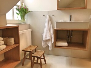Un baño con un estante de madera que sostiene toallas, un taburete pequeño, una toalla blanca colgada en un gancho y un espejo sobre el lavabo.