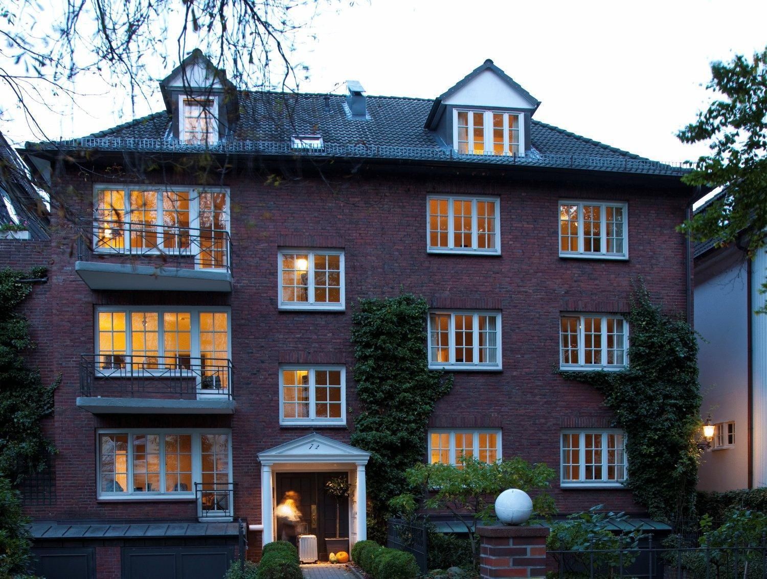 Ein mehrstöckiges Backsteingebäude mit einer symmetrischen Fassade, großen Fenstern und einem zentralen Eingang mit einer Tür und einem kleinen Vorbau.