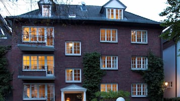 Ein mehrstöckiges Backsteingebäude mit einer symmetrischen Fassade, großen Fenstern und einem zentralen Eingang mit einer Tür und einem kleinen Vorbau.