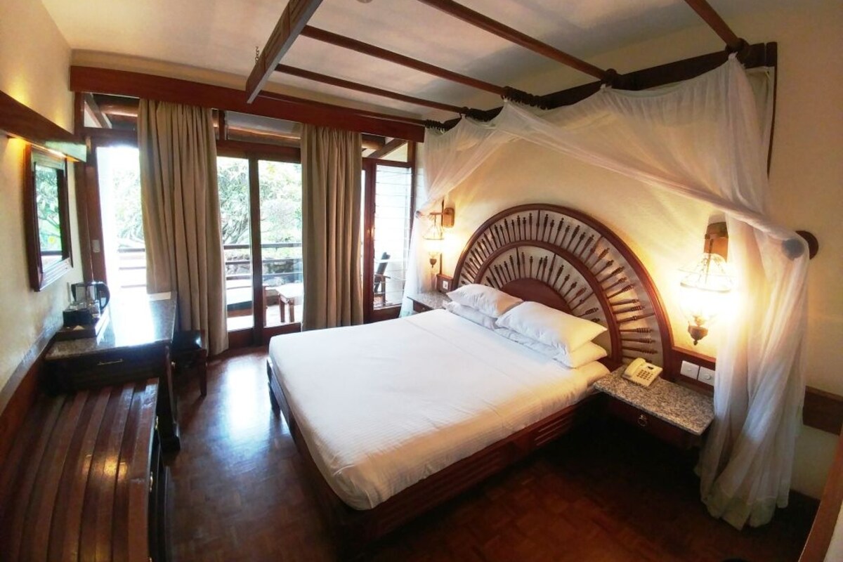 Une chambre avec un grand lit, un plancher en bois, un lit à baldaquin, une table de chevet avec une lampe et une vue sur l’extérieur.