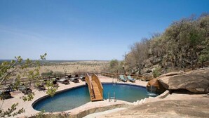 A pool with a wooden slide, lounge chairs, and a rocky landscape in the background.