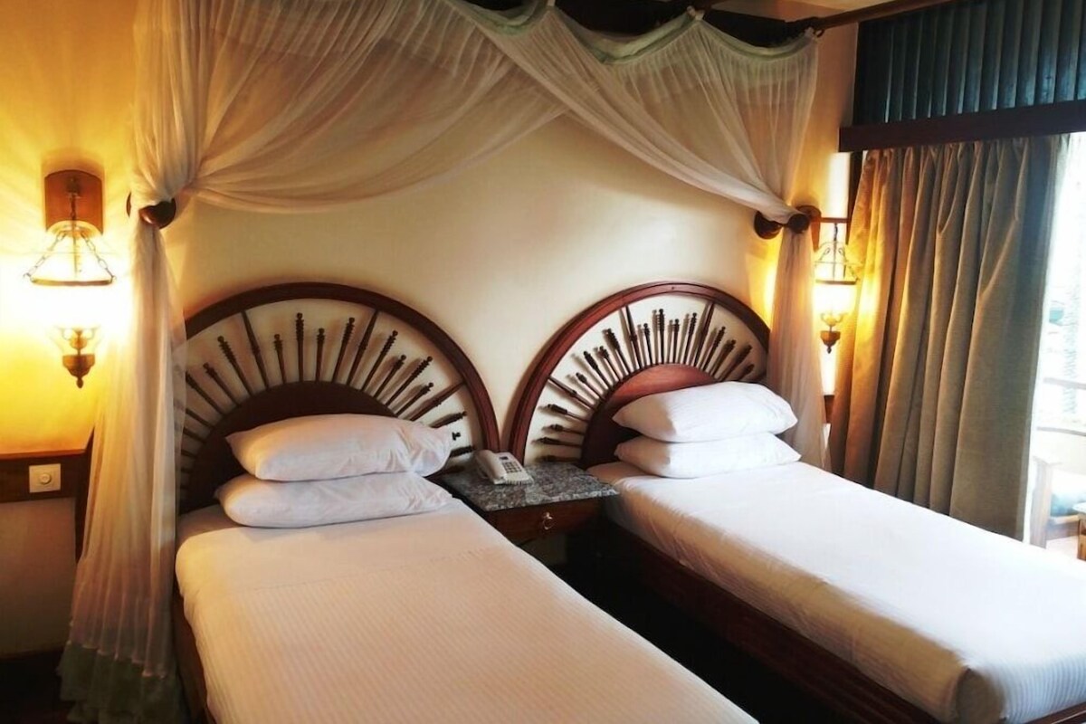 A hotel room with two beds, each with a canopy, bedside lamps, and a small table between the beds.