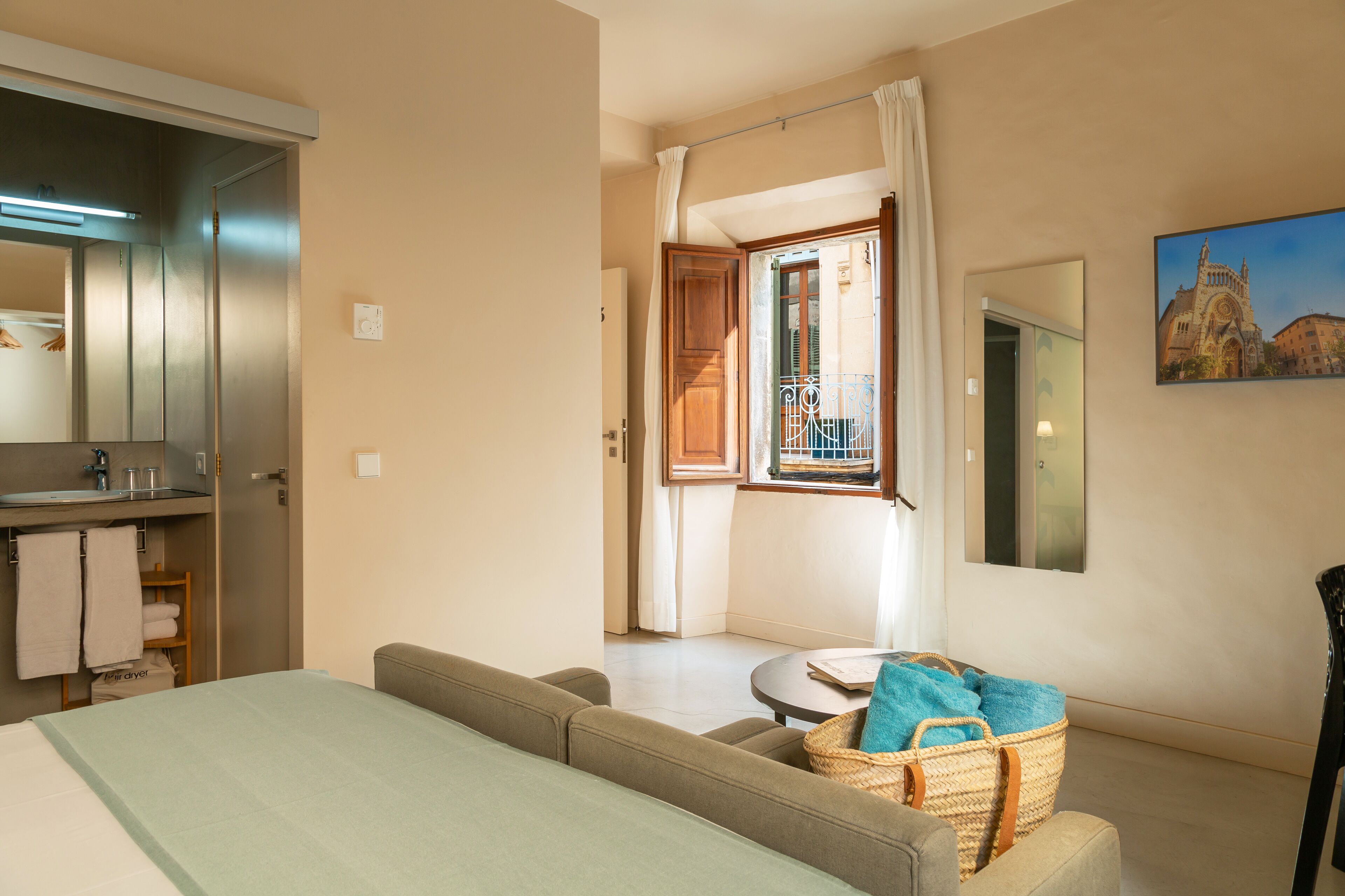 A hotel room with a bed, a sofa, a small round table, a wicker basket, a mirror, and a painting of a building.