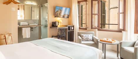 A hotel room with a bed, a desk, a chair, a small table with a tray, a TV, and a window with shutters.