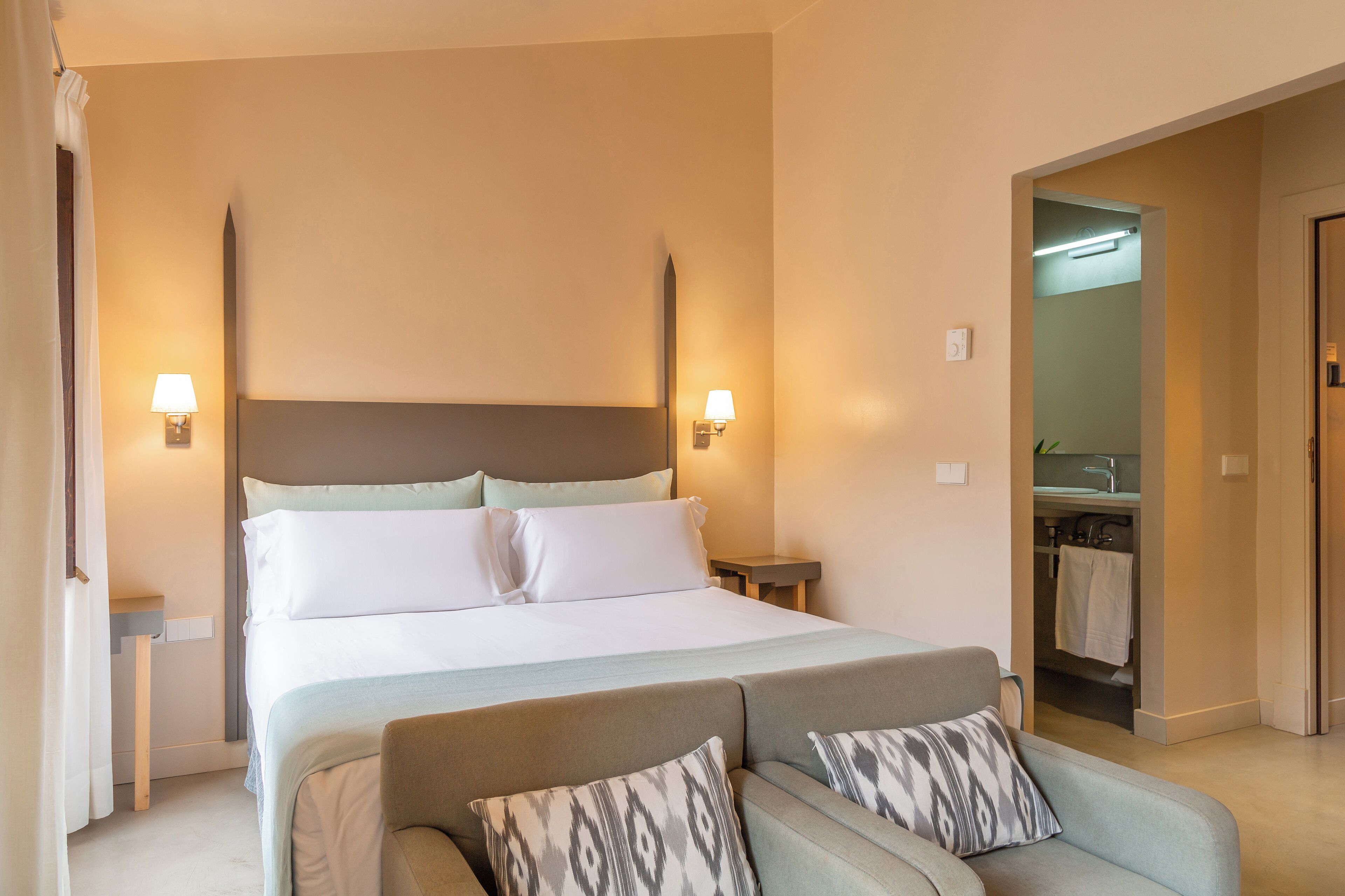 A hotel room with a large bed, two bedside tables with lamps, a sofa with patterned cushions, and a bathroom visible through an open door.