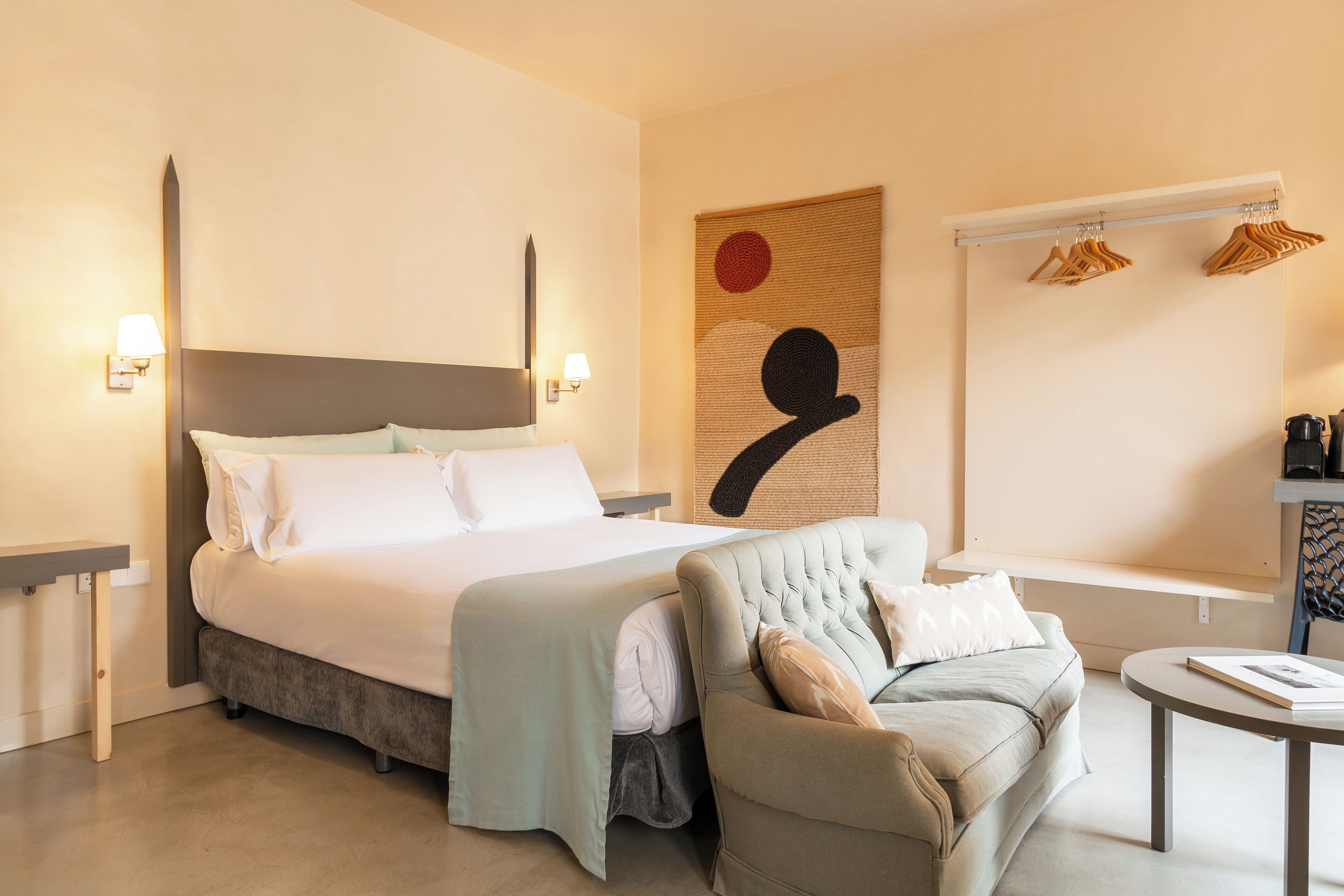 A modern hotel room with a large bed, a sofa, a small table, and a wall-mounted coat rack.