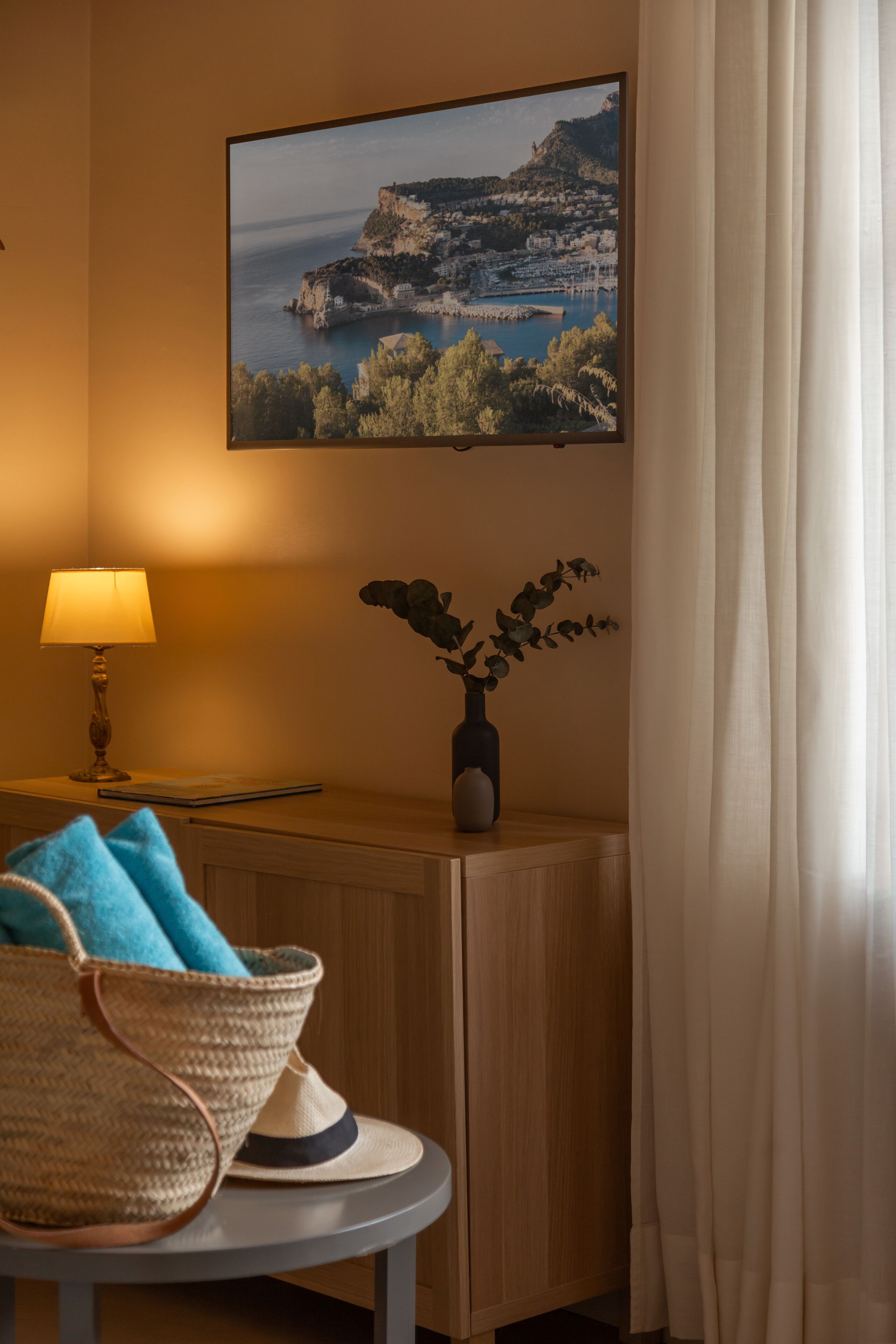 A room with a wooden cabinet, a framed landscape painting, a lamp, a vase with a plant, and a wicker chair with a blue towel.