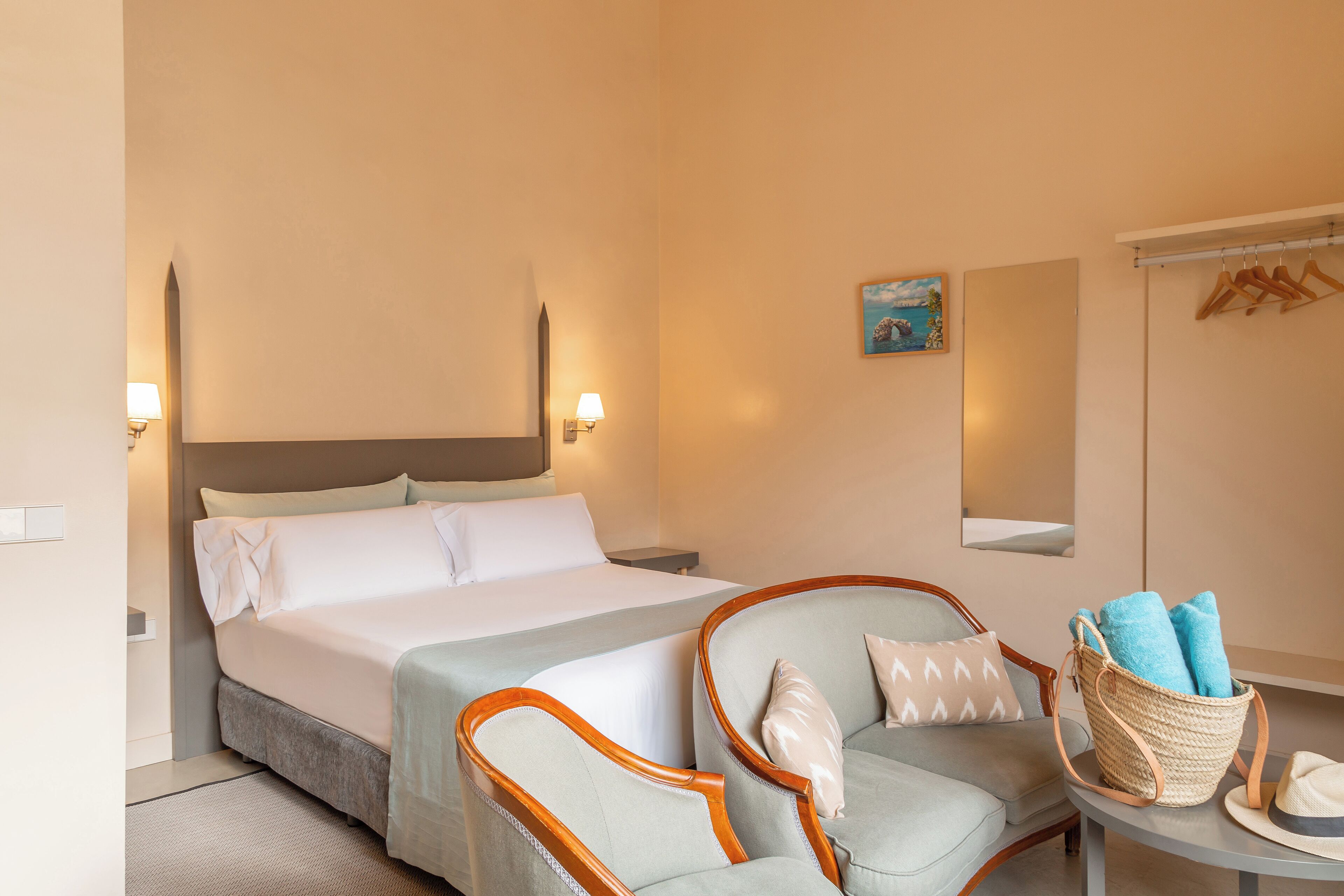 A hotel room with a bed, two armchairs, a small table with a basket of towels, and a wall-mounted mirror.