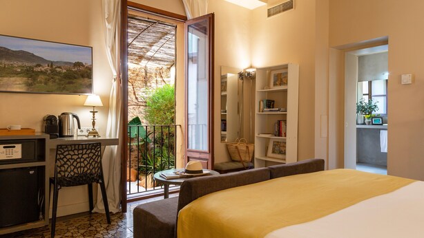 A hotel room with a bed, a desk, a chair, a small table, a bookshelf, and a balcony with a view of greenery.
