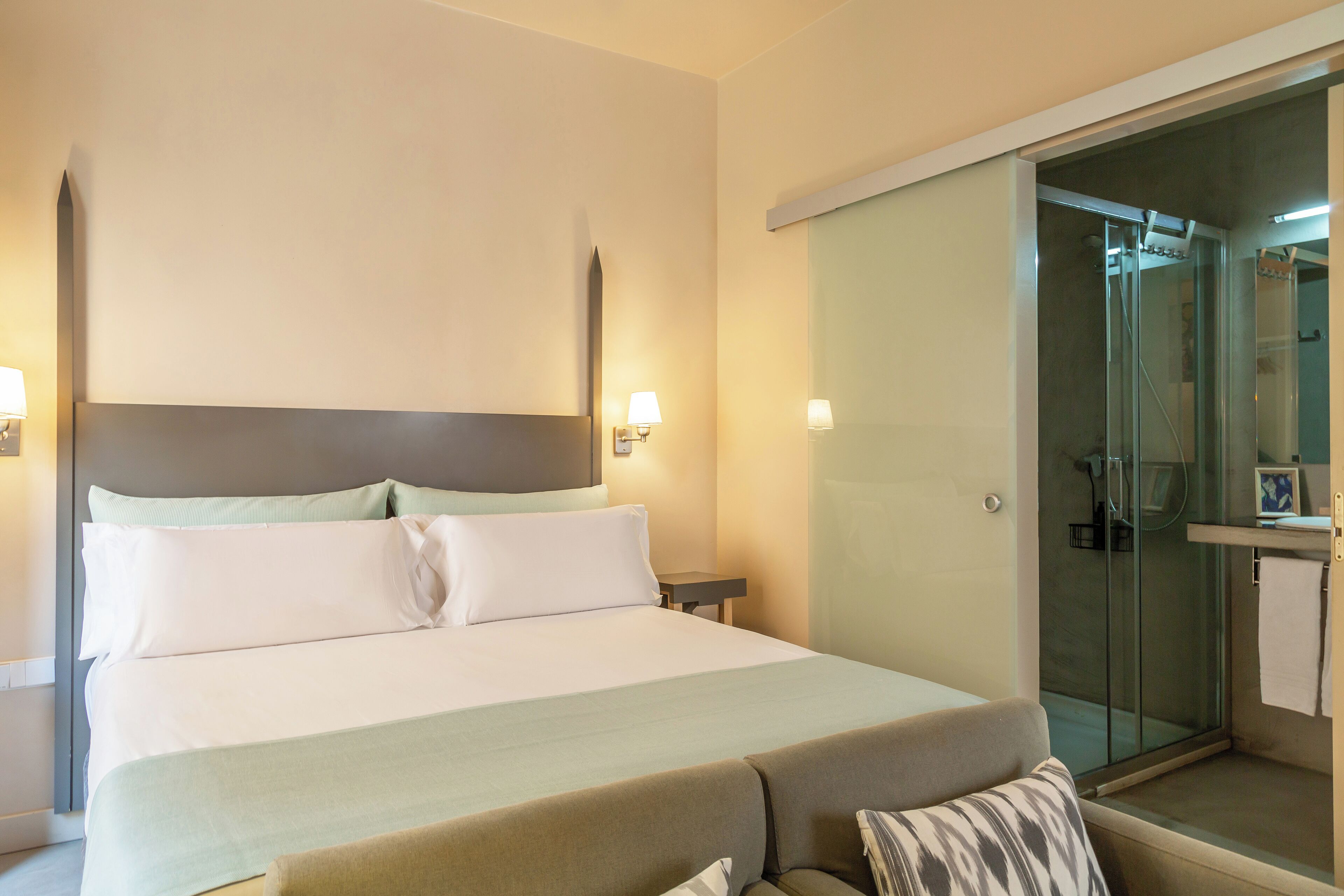 Une chambre d’hôtel dotée d’un grand lit, d’une table de chevet, d’une petite commode, d’une douche en verre et d’un petit coin salon.
