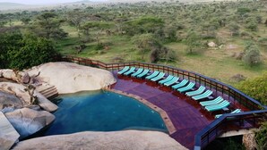A pool with lounge chairs, surrounded by a wooden deck and a stone wall, overlooking a savannah landscape. - Seronera Wildlife Lodge (Serengeti)
