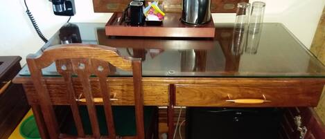 A wooden dining table with a glass top, a chair, a small black appliance, and a mirror reflecting a person.