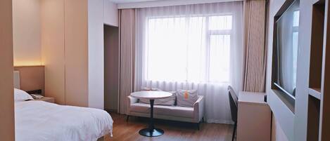 A hotel room with a bed, a small round table, a sofa, a TV, and a window with curtains.