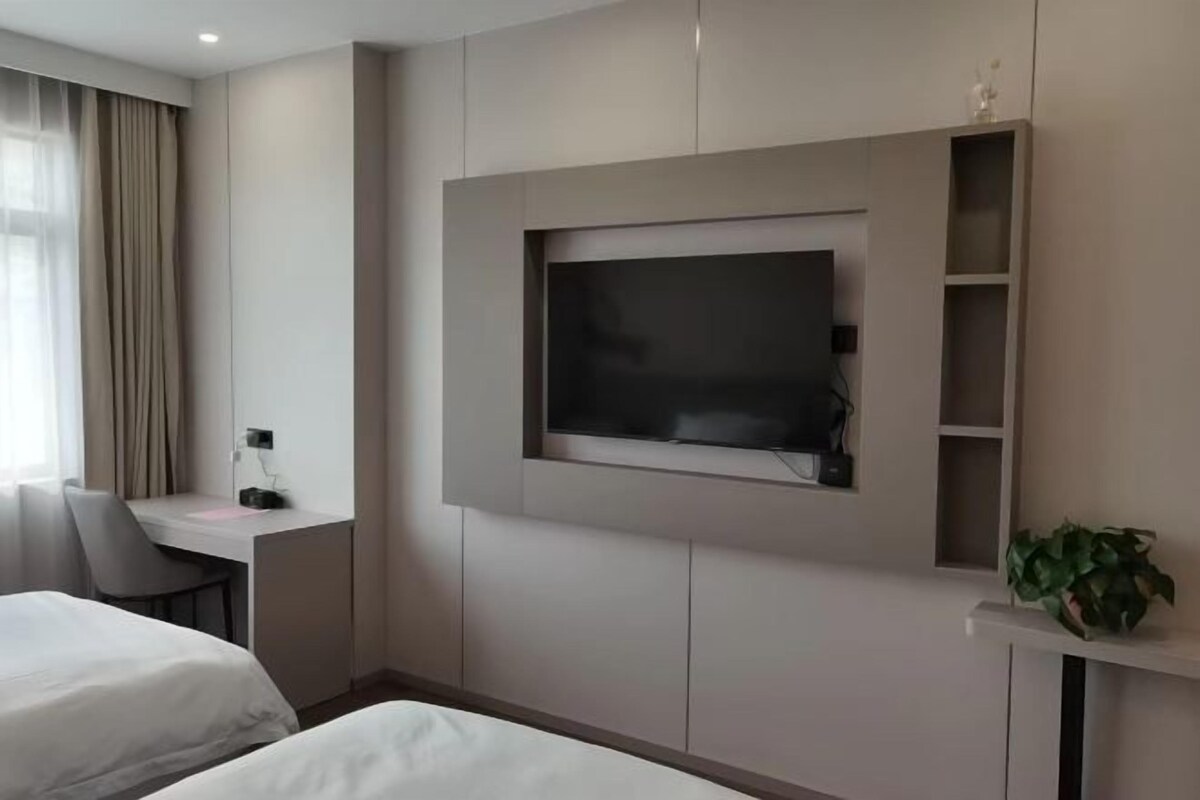 A hotel room with a bed, a desk, a TV mounted on the wall, and a potted plant.