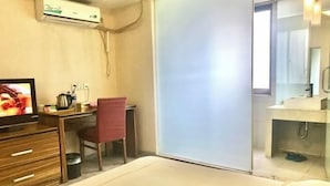 Quarto de hotel com cama, escrivaninha com cadeira, televisão, espelho e banheiro com pia e chuveiro.