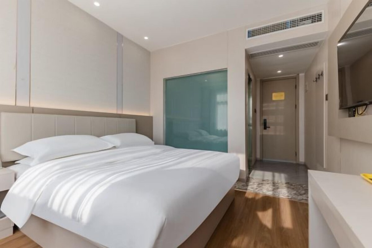 A modern hotel room with a large bed, a bedside table, a TV, and a bathroom visible through a glass door.