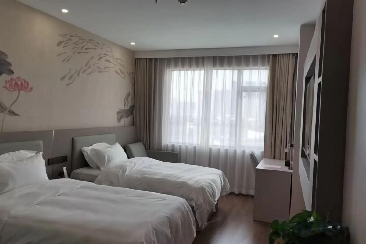 A hotel room with two beds, a small bedside table, a window with curtains, and a wall with floral artwork.