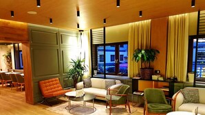 A modern living room with a sofa, armchairs, and a coffee table. There are potted plants and a view of the city through large windows. - St. Joseph Hotel (Hamburg)