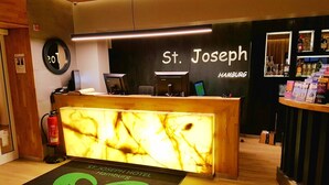 A reception desk at St. Joseph Hotel Hamburg with a marble front, a fire extinguisher, a reception area, and a welcome mat. - St. Joseph Hotel (Hamburg)