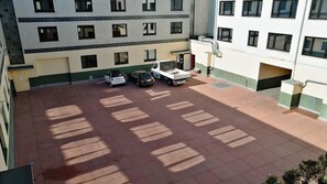 Un immeuble à plusieurs étages doté d’un espace de stationnement. Plusieurs voitures y sont garées, dont un fourgon blanc et une voiture noire. L’immeuble possède de nombreuses fenêtres et une façade aux couleurs verte et blanche.