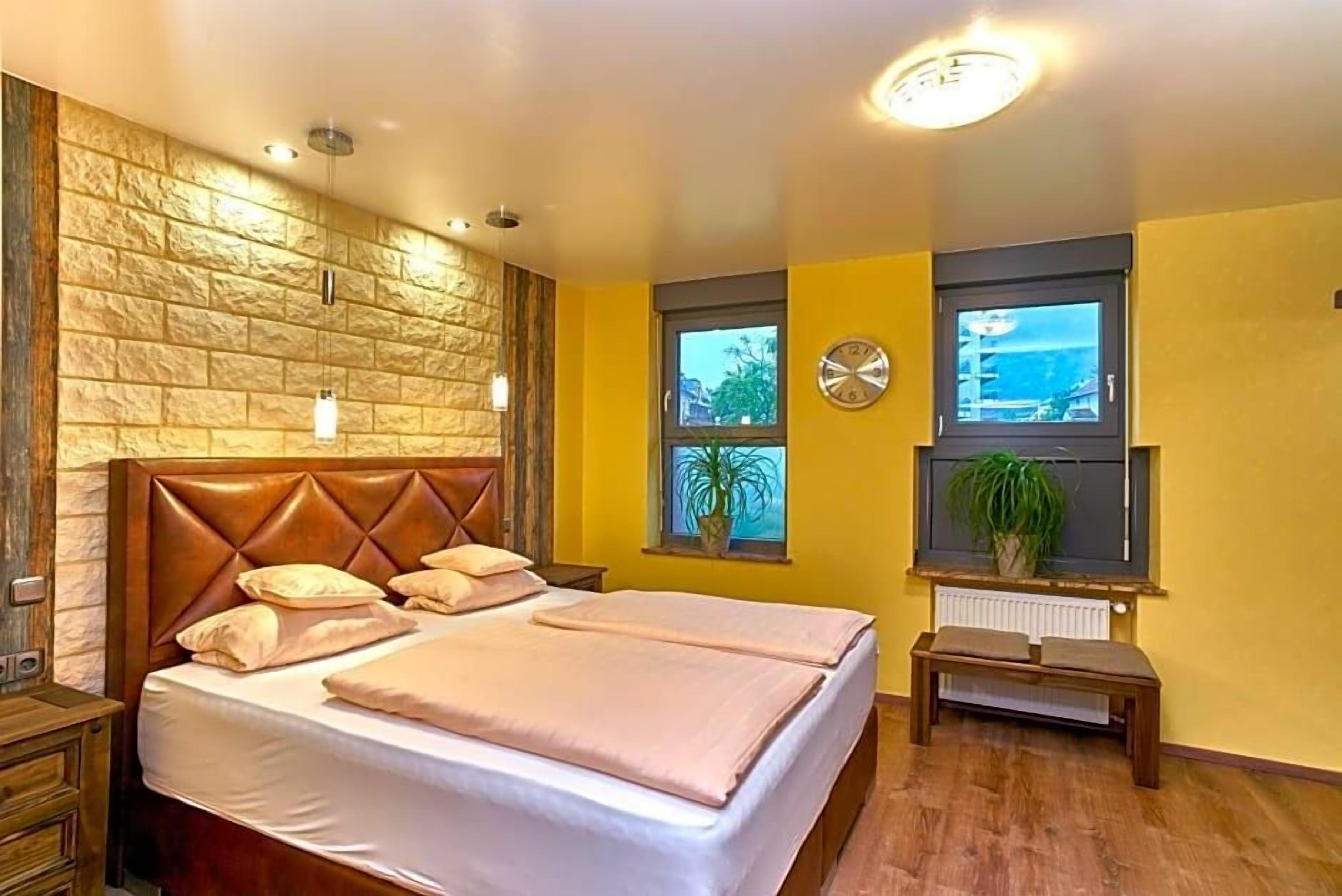 Une chambre à coucher avec une tête de lit ornée d’un motif en briques, deux fenêtres avec des plantes, une table de chevet et un banc.