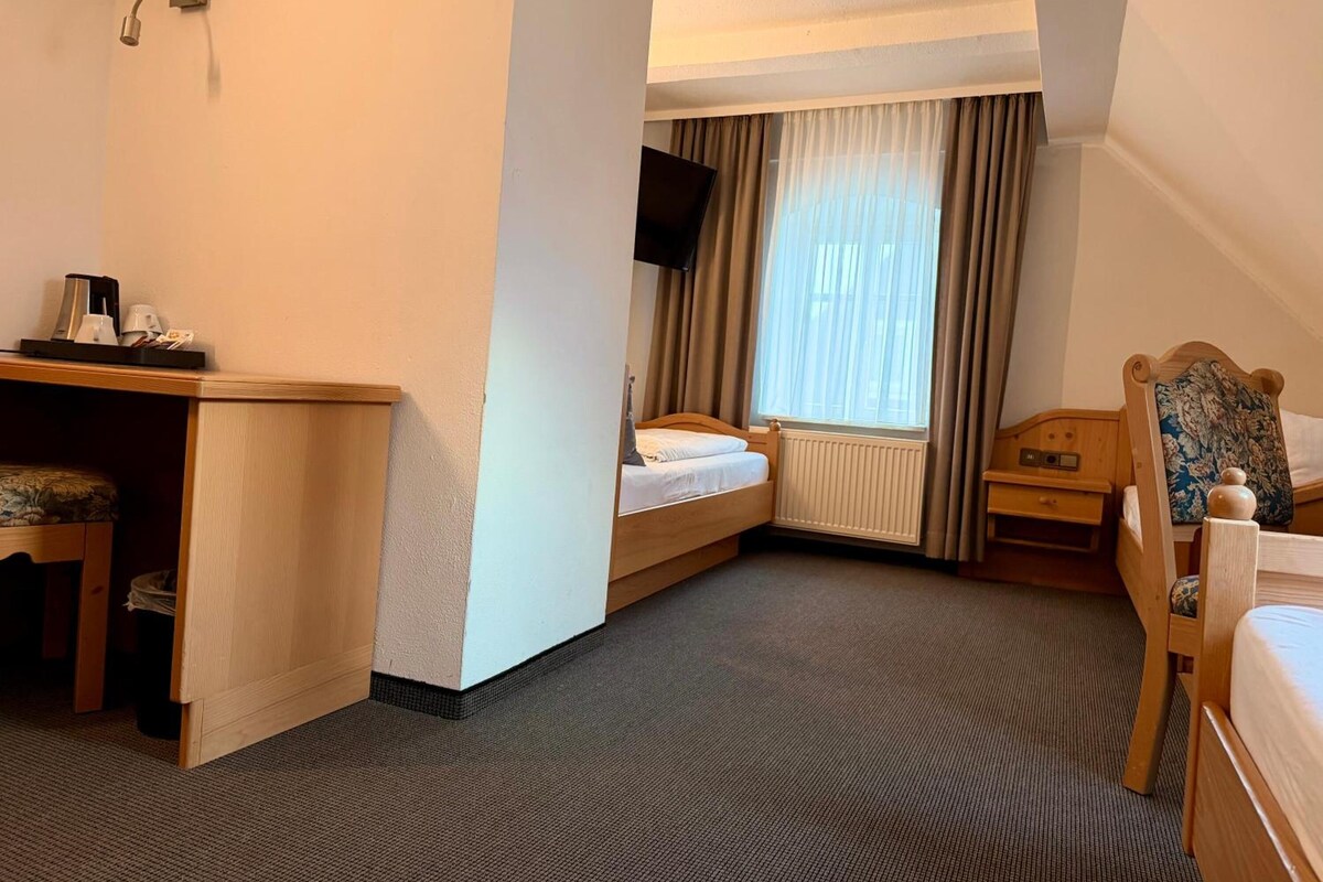 Quarto de hotel com cama, escrivaninha, cadeira, televisão e janela com cortinas.