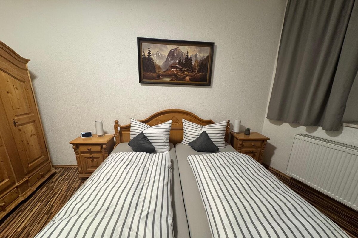 Uma cama de casal com roupa de cama listrada, cabeceira de madeira, duas mesas de cabeceira com luminárias, um quadro na parede, um guarda-roupa de madeira e um radiador.