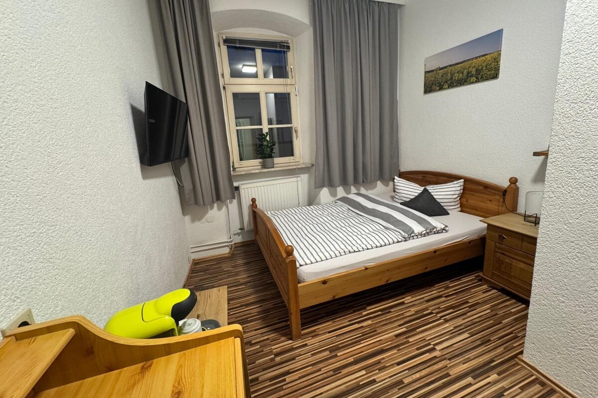 Quarto com cama de madeira, mesa de cabeceira pequena, cadeira, janela com cortinas e TV fixada na parede.