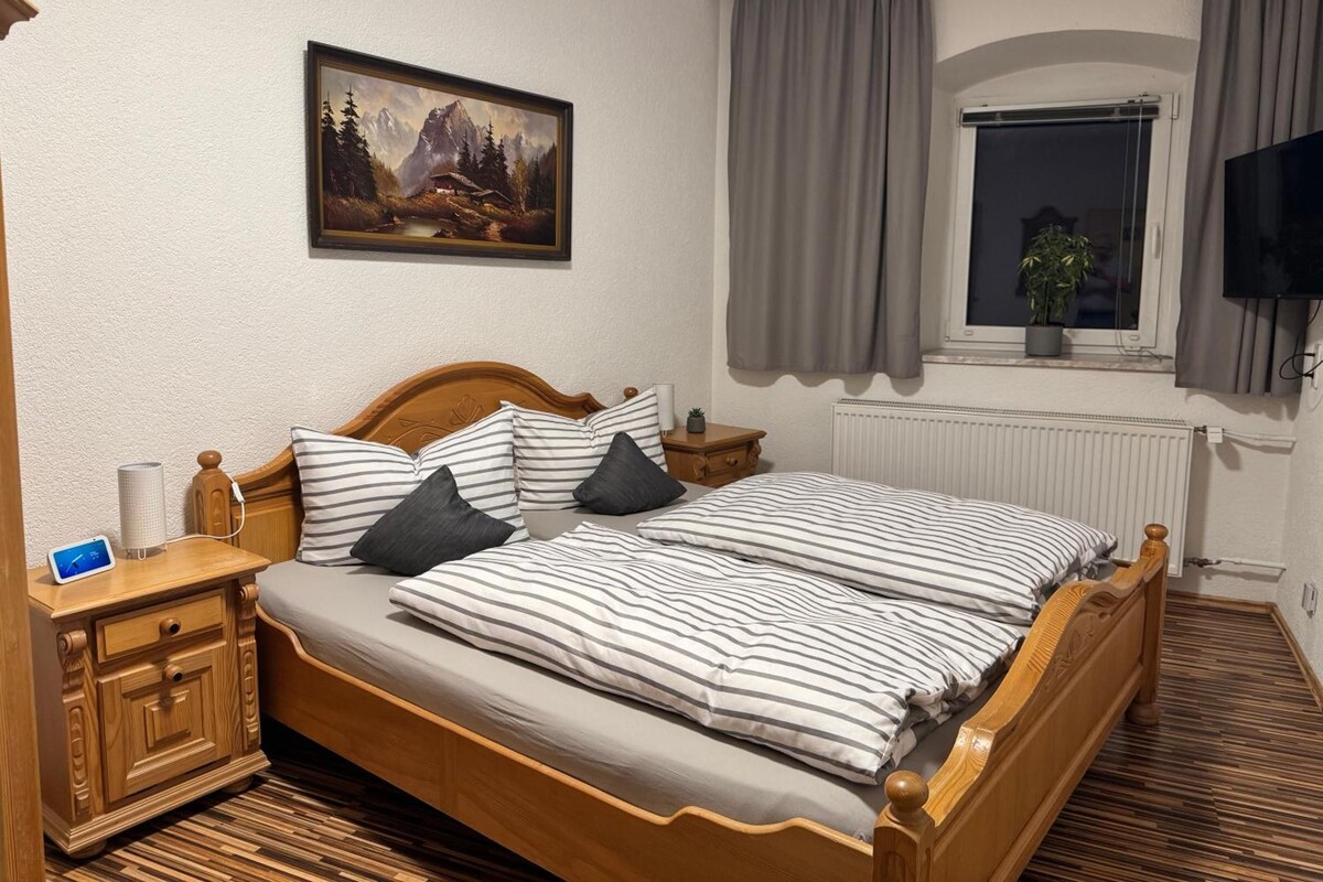 A double bed with striped bedding, a nightstand with a lamp, a painting of a mountain scene, a window with curtains, and a television.