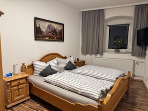 A double bed with striped bedding, a nightstand with a lamp, a painting of a mountain scene, a window with curtains, and a television.