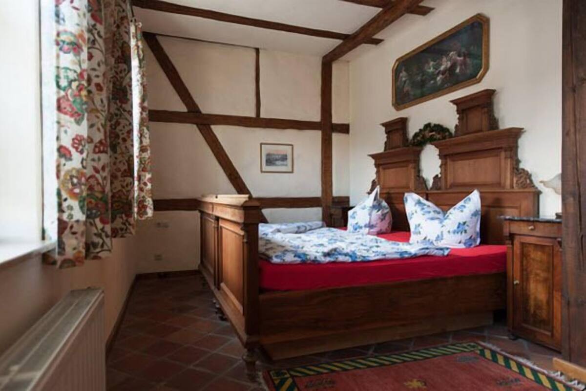 Un dormitorio con cama de madera, cortinas florales, colcha roja y un cuadro en la pared.