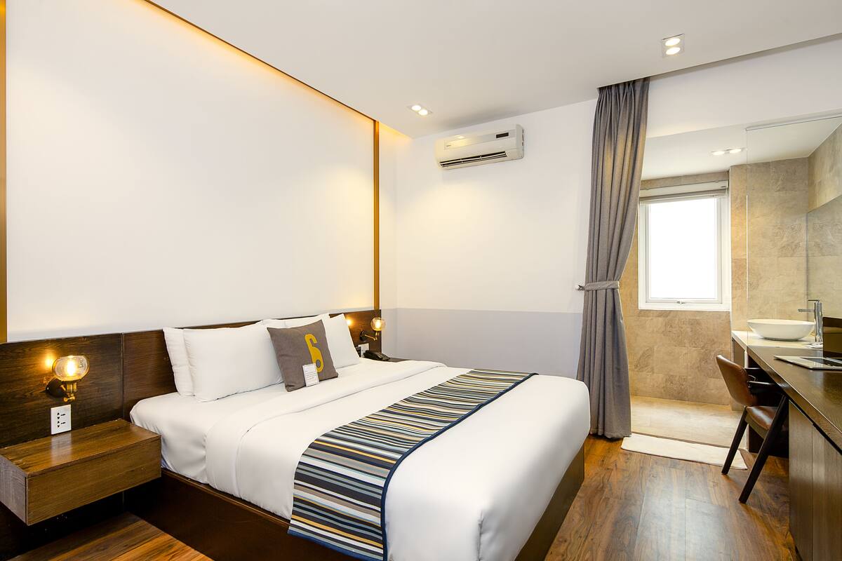 Una habitación de hotel moderna con una cama grande, mesita de noche y un baño con lavamanos y espejo.