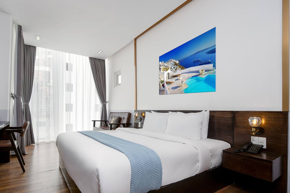 Una habitación de hotel moderna con una cama grande, suelo de madera, un escritorio con una silla, una mesita de noche y una lámpara de pared.