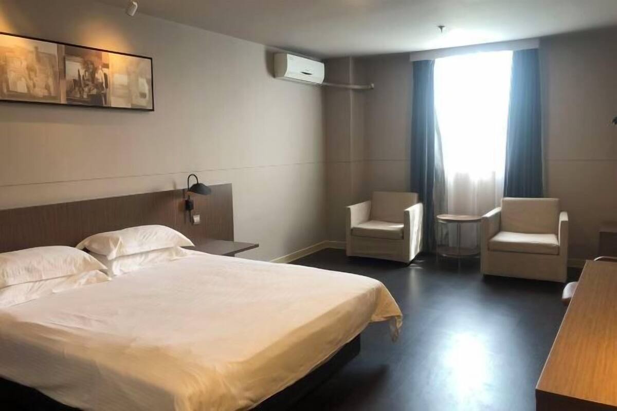 Une chambre d’hôtel avec un lit, deux chaises, une petite table, un appareil de climatisation et un tableau au mur.