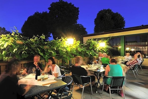 Un área de comedor al aire libre con mesas y sillas, rodeada de follaje y un edificio al fondo.