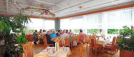 A dining area with multiple tables and chairs, large windows, and indoor plants.