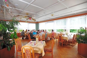 Un comedor con varias mesas y sillas, ventanas amplias y plantas de interior.