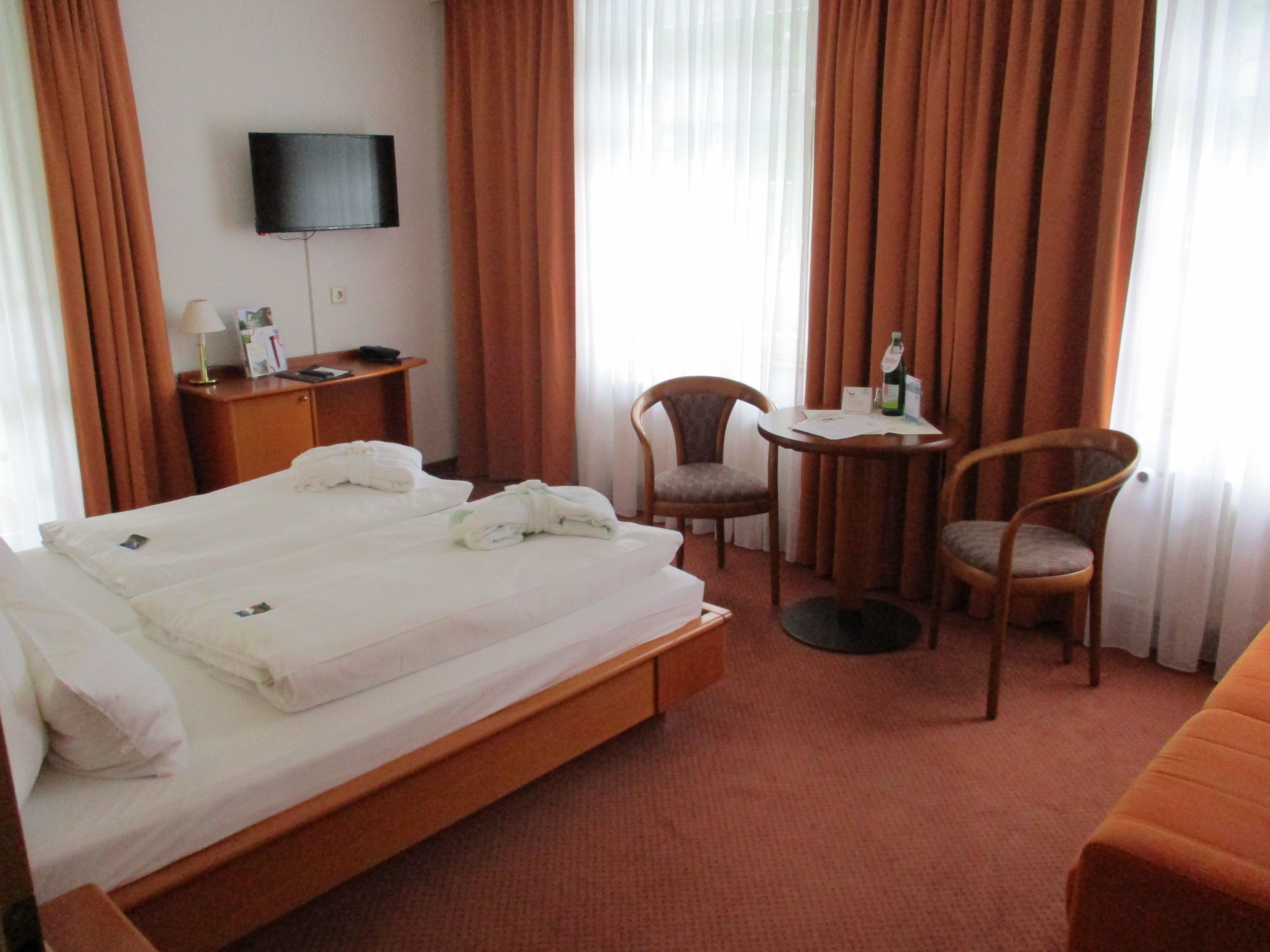 A hotel room with a bed, two chairs, a small table, a TV, and a desk with a lamp and a bottle.