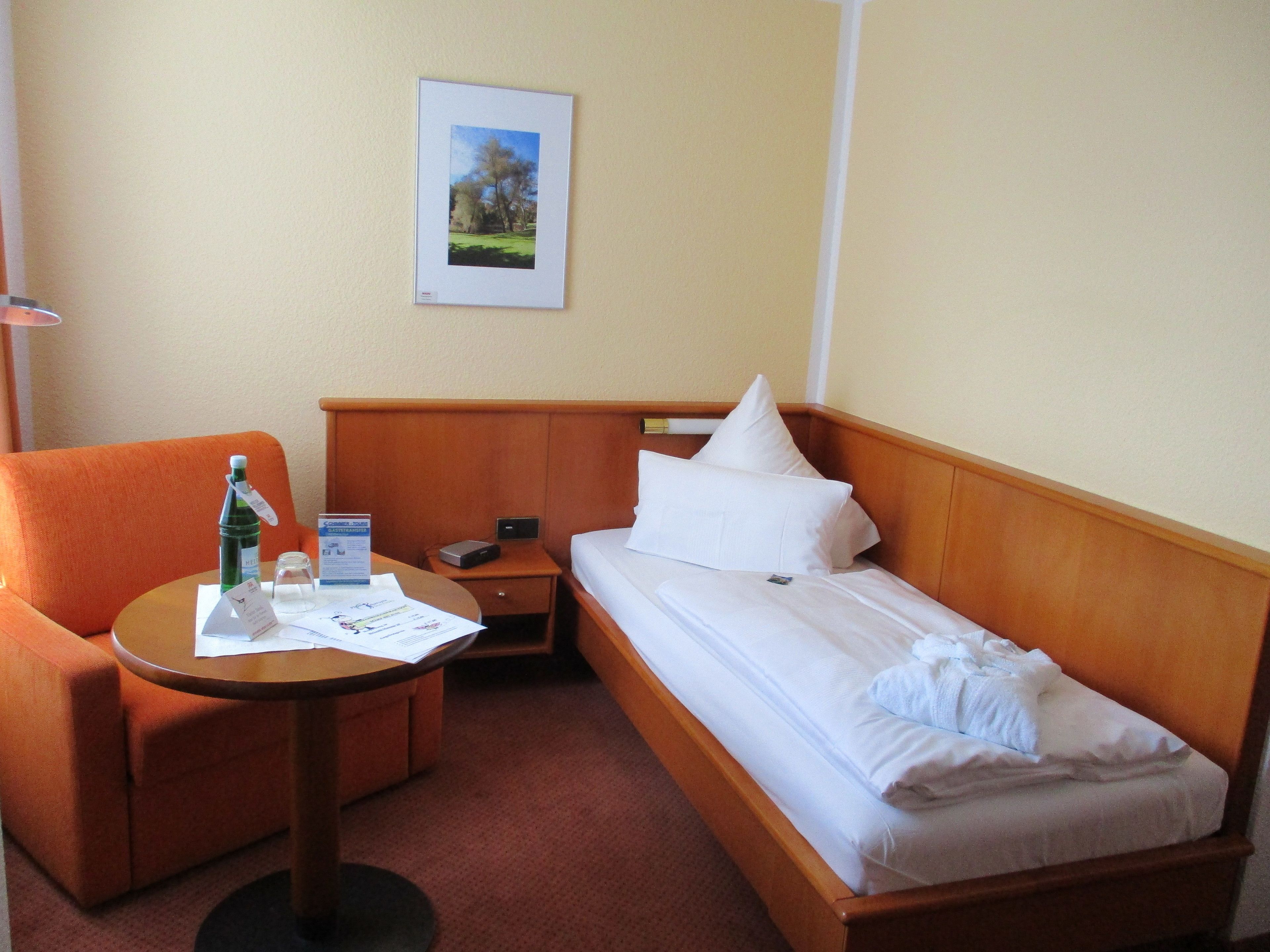 A hotel room with a single bed, a small table with a bottle, a chair, a nightstand, and a framed picture on the wall.