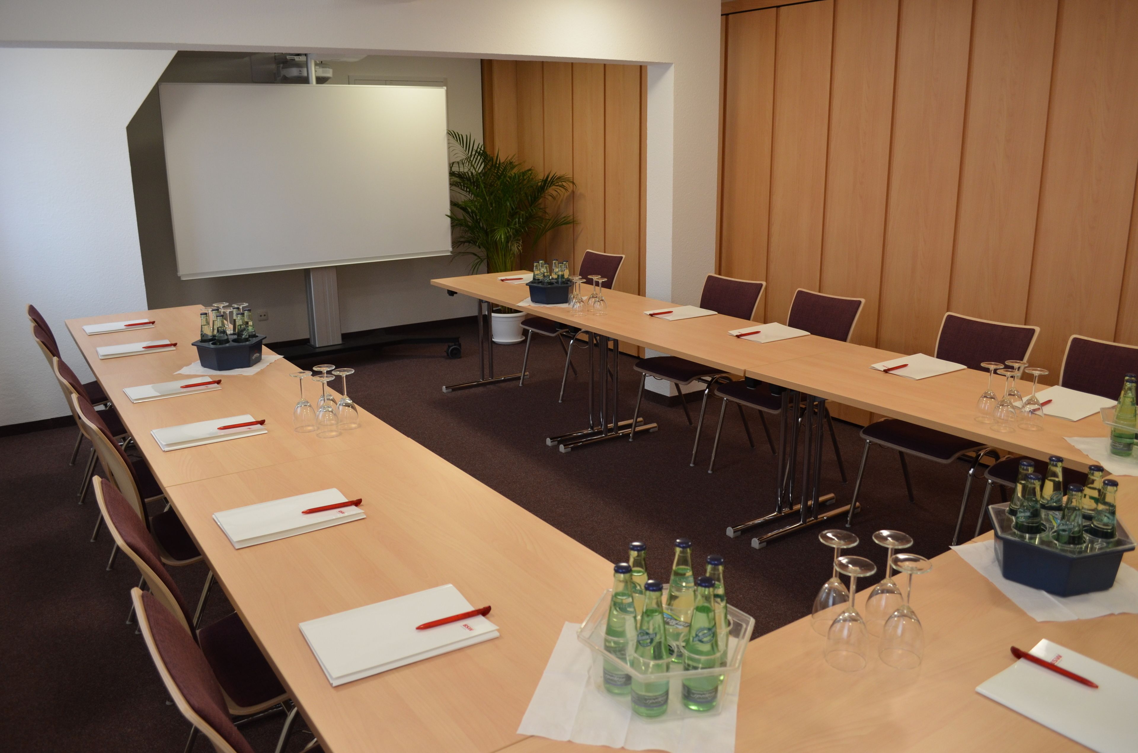 A conference room with a long table, chairs, a projector screen, and a potted plant.