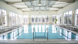 Una piscina interior con paredes de vidrio transparente, sillas reclinables blancas y vistas a árboles al exterior.