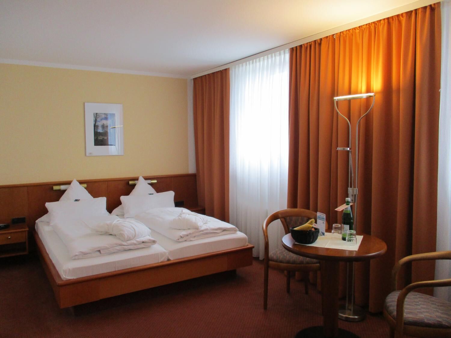 A hotel room with a double bed, a bedside table, a chair, a small table with a bottle and glass, and a window with curtains.