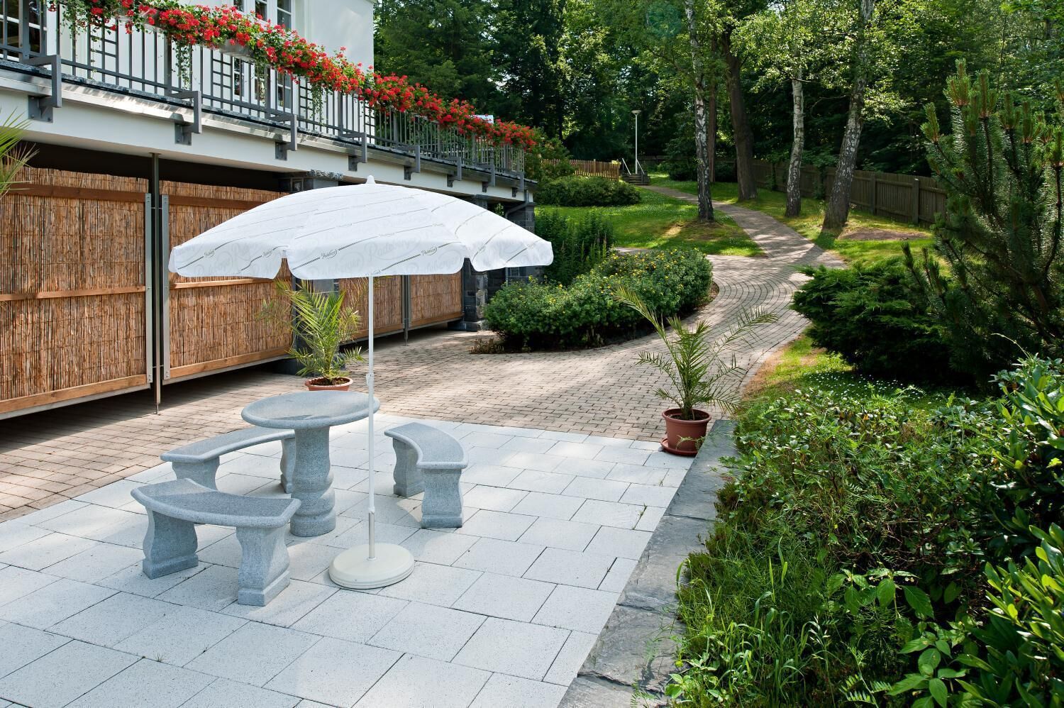 Een terras buiten met stenen tafels en banken, omringd door weelderige begroeiing en een houten omheining.