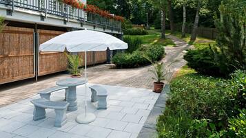 Een terras buiten met stenen tafels en banken, omringd door weelderige begroeiing en een houten omheining.