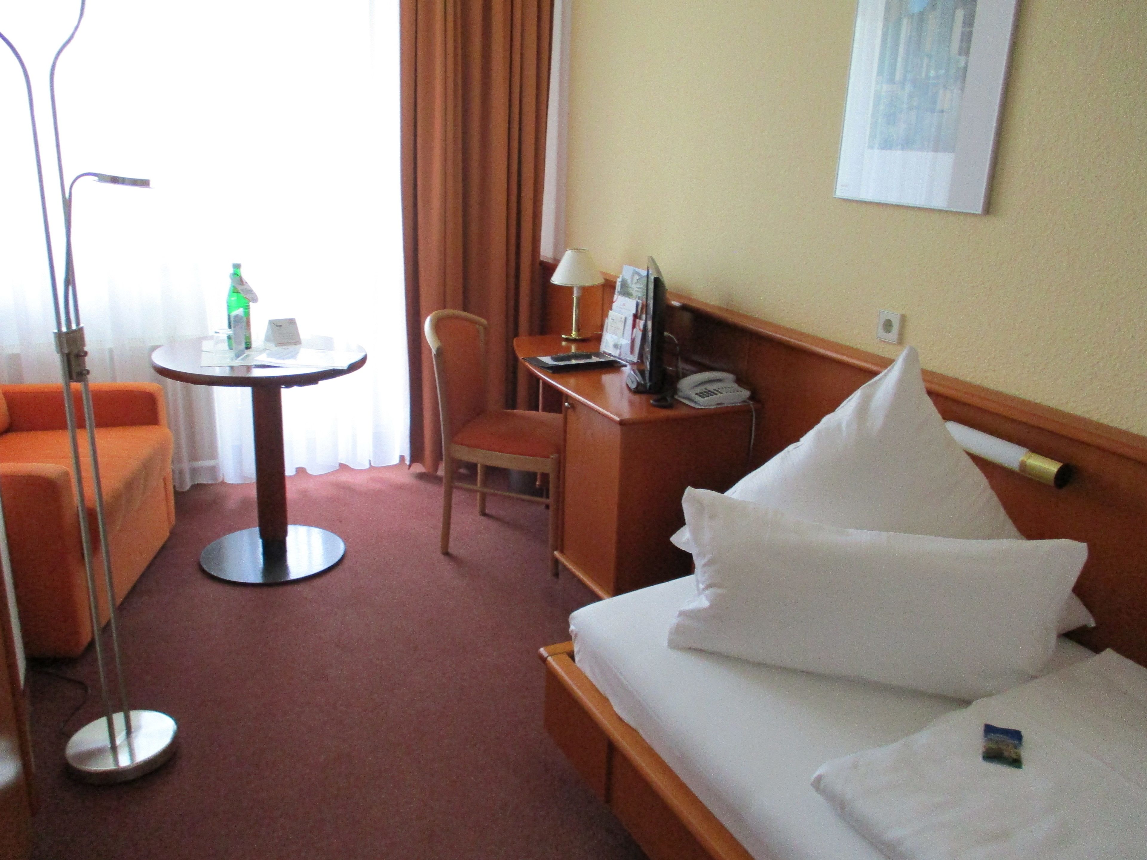 A hotel room with a bed, a desk, a chair, a table, and a lamp.
