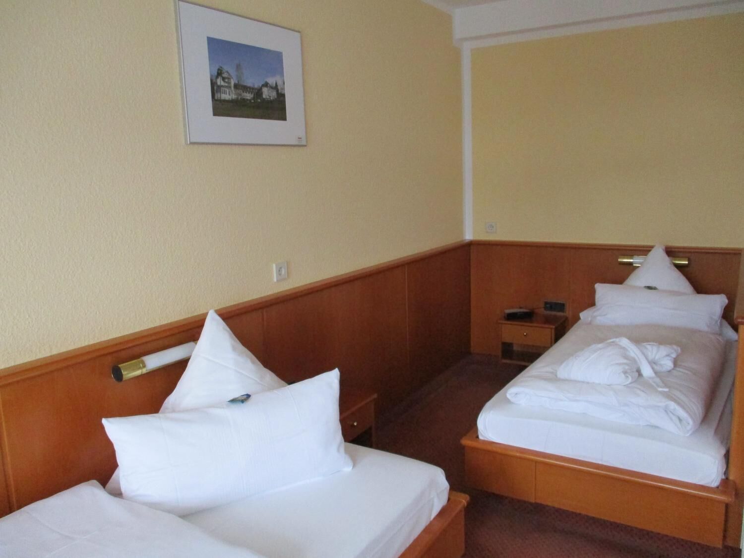 A hotel room with two single beds, wooden headboards, a small bedside table, and a framed picture on the wall.