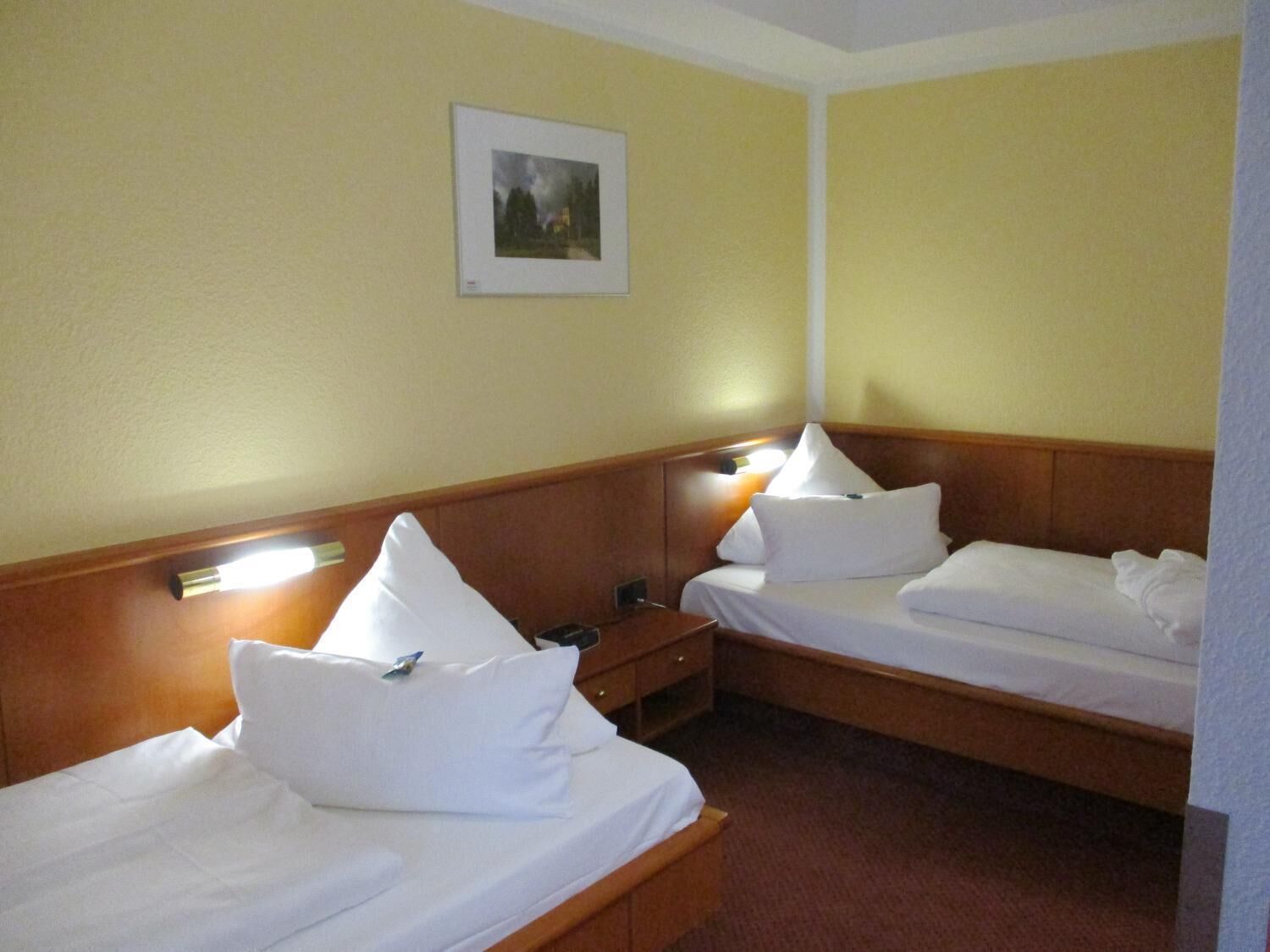 Een hotelkamer met twee bedden, elk voorzien van wit beddengoed en kussens, een nachtkastje en een ingelijst schilderij aan de muur.