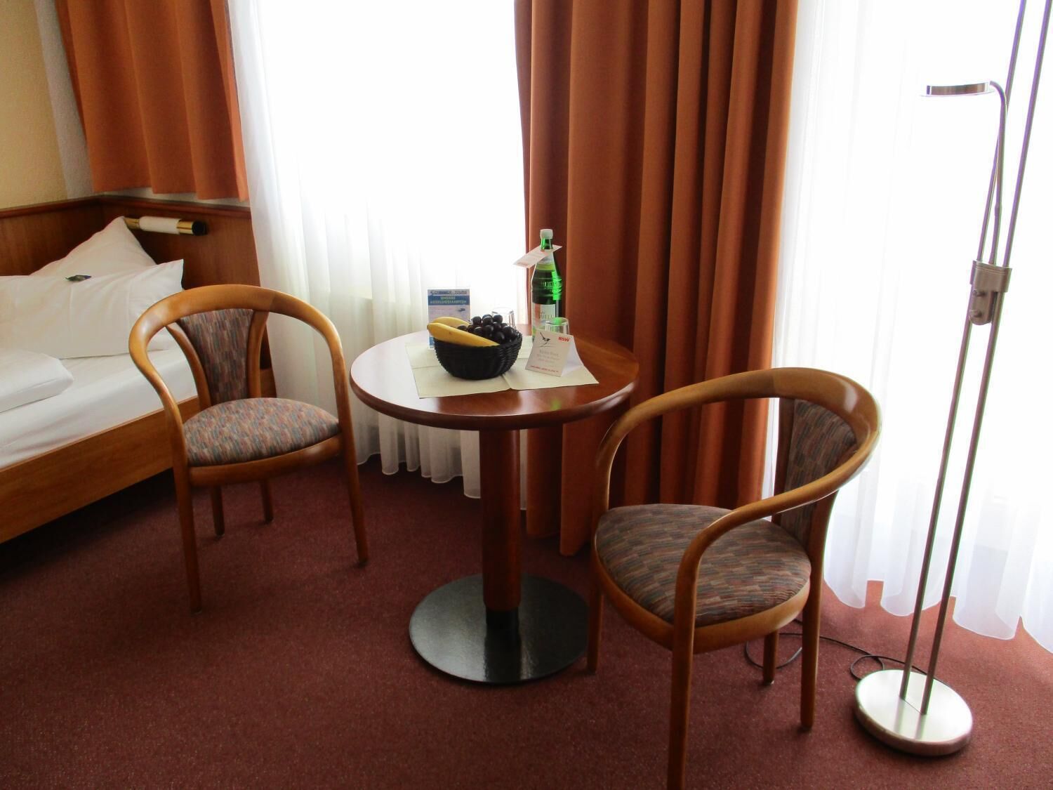 A hotel room with a bed, two armchairs, a small round table with a bowl of fruit and a bottle, and a floor lamp.