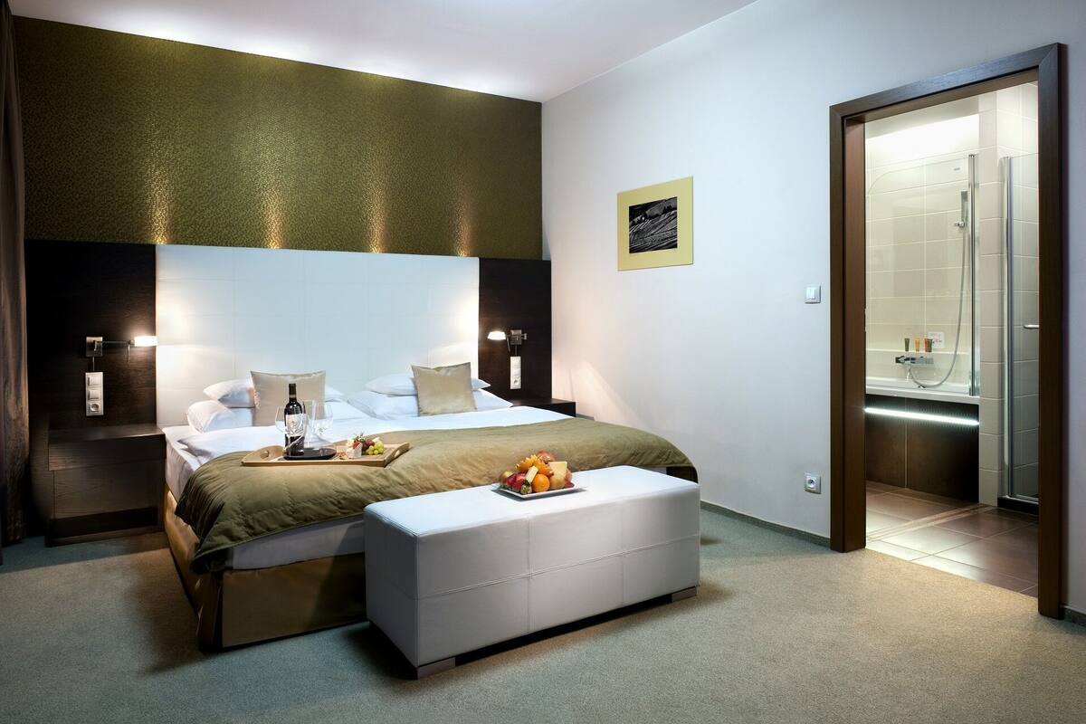 Quarto de hotel moderno com uma cama grande, mesas de cabeceira, uma pequena mesa com frutas e uma garrafa, um banco, um banheiro com chuveiro e um quadro na parede.